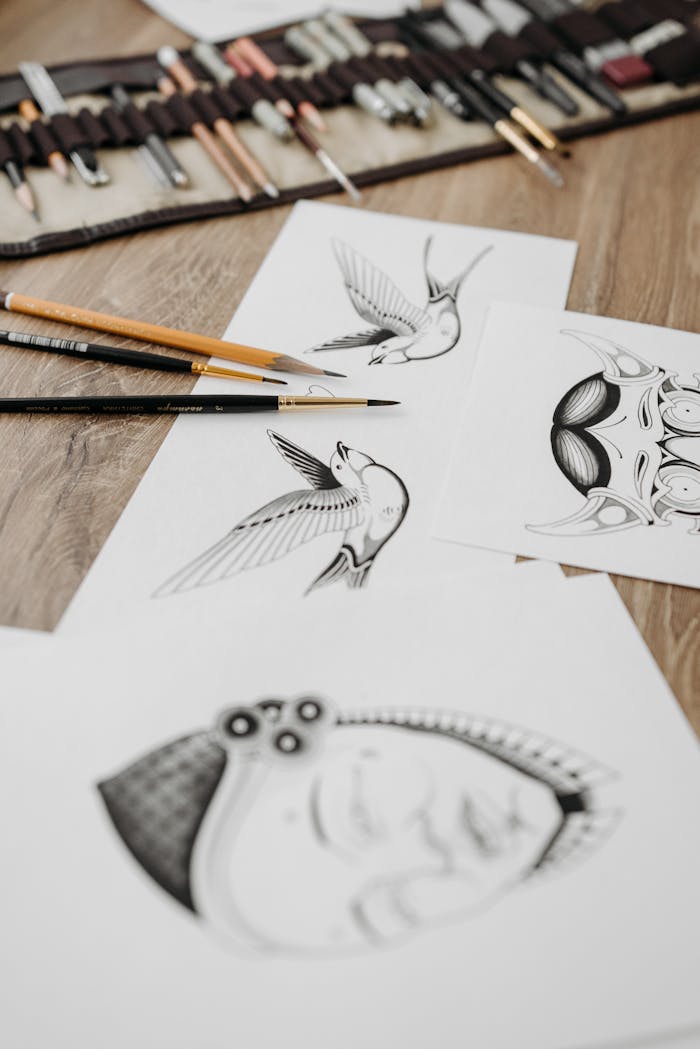 services-02 Detailed pencil sketches of tattoos with artistic designs on a wooden surface. Includes tools like paintbrushes and pencils.